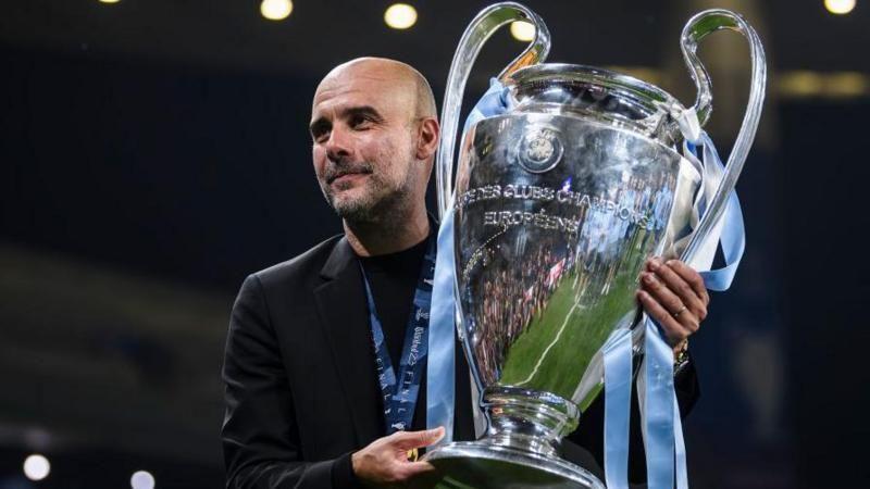 المدرب الإسباني بيب غوارديولا يحمل كأس دوري أبطال أوروبا - فاز به فريقه مانشستر سيتي 2023
يرتدي سترة سوداء ويحمل كأسا فضية تتدلى منها شرائط زرقاء - ويضع ميدالية ذهبية