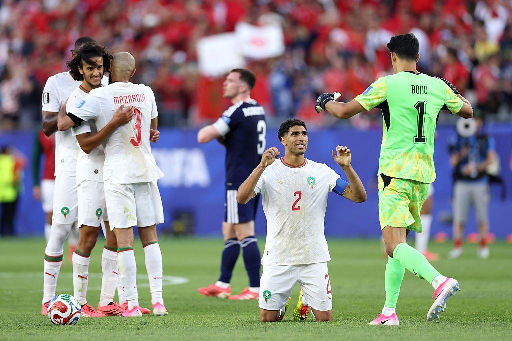 لاعبو المنتخب المغربي، ومن بينهم قائد الفريق أشرف حكيمي وحارس المرمى ياسين بونو، يحتفلون عقب انتهاء مباراتهم أمام اسكتلندا، فيما يظهر في الخلفية قائد المنتخب الاسكتلندي أندي روبرتسون، وقد بدا عليه الحزن، بينما تحتفل الجماهير المغربية بالفوز.