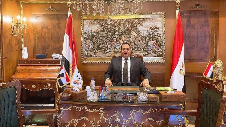 صبري نخنوخ يجلس على كرسي وأمامه مكتب خشبي مزكرش بلون ذهبي وحوله أعلام مصر وخلفه لوحة، ويبدو أن أثاث المكتب ثمين. وفي السقف إضاءة كريستالية كبيرة. يرتدي بدلة رسمية تميل إلى اللون البني مع قميص أبيض وربطة عنق بنفس لون البدلة منطقة كما يبدو باللون بالذهبي.