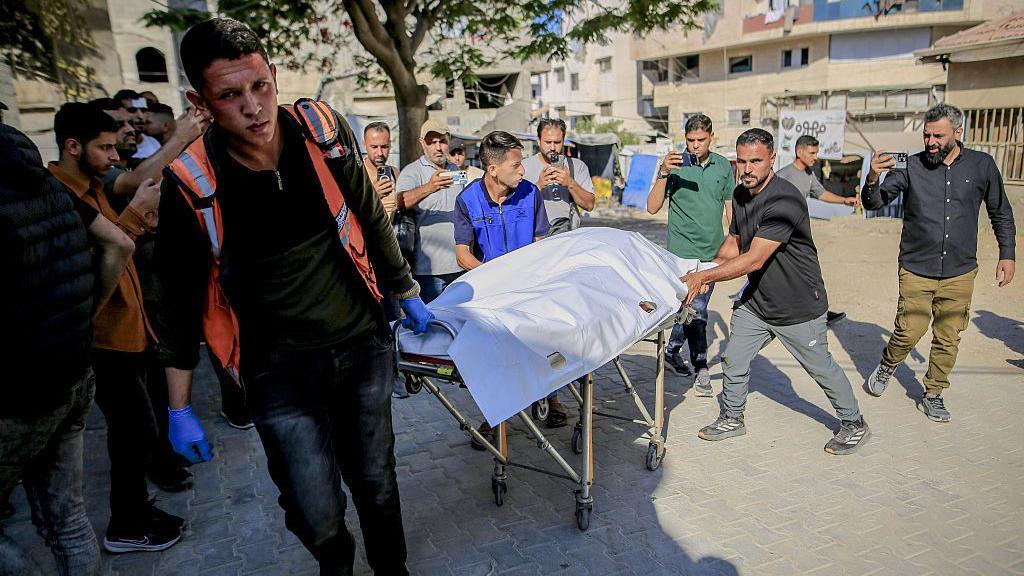 نقل جثامين ضحايا فلسطينيين سقطوا في هجوم إسرائيلي إلى مستشفى الشفاء لإجراء مراسم الدفن في مدينة غزة، يظهر في الصورة الملتقطة نهاراً مسعف وشخصان يجرون عربة عليها جثمان ملفوف بالأبيض وحولهم عدد من الأشخاص.